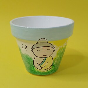 May include: A small terracotta flower pot painted with a cartoon image of a meditating Buddha in a yellow robe. The pot is painted with green grass and a yellow sun.