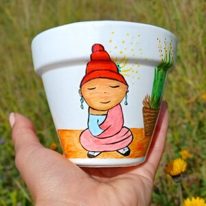 Può includere: Un vaso di ceramica bianca con un disegno dipinto a mano di una persona che indossa un cappello rosso e una veste rosa. La persona sta meditando a occhi chiusi. Il vaso ha uno sfondo giallo, arancione e verde.