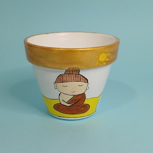 May include: White ceramic planter with a gold rim and a painted image of a meditating Buddha with a yellow background and a sun above the Buddha.