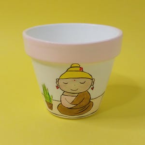 May include: A white ceramic planter with a pink rim, featuring a hand-painted illustration of a meditating Buddha with a yellow robe and closed eyes. The Buddha is surrounded by green plants.