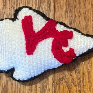 May include: A white crocheted arrowhead with a red crocheted "K" in the center. The arrowhead has a black crocheted outline.