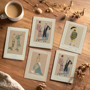 May include: A collection of vintage fashion illustration prints featuring women in stylish dresses and coats. The prints are in a variety of colors, including pink, black, and green, and are arranged on a wooden surface with a cup of coffee.
