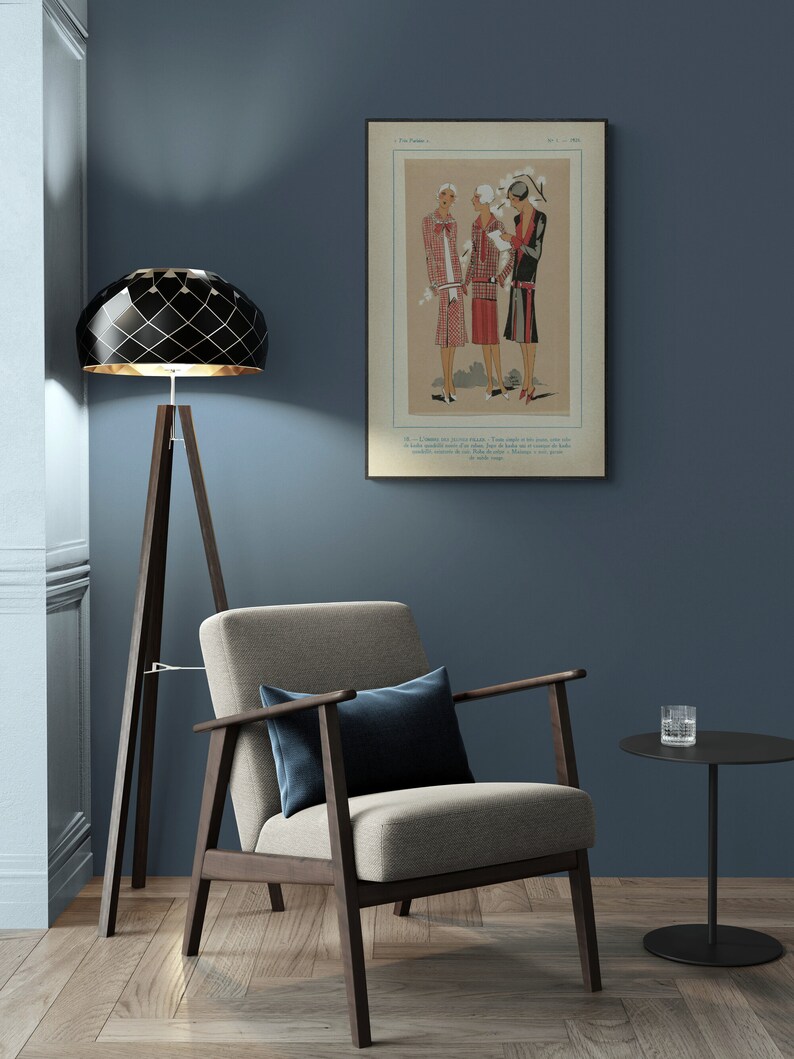 Pode incluir: Um candeeiro de ch&atilde;o geom&eacute;trico preto e dourado com uma base de trip&eacute;, uma poltrona bege com um almofada azul escuro e uma mesa auxiliar preta com um copo em cima. A parede &eacute; de cor azul escura e uma ilustra&ccedil;&atilde;o vintage emoldurada de tr&ecirc;s mulheres em vestidos est&aacute; pendurada na parede.