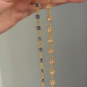 May include: A pair of gold-colored earrings with a floral design. Each earring features alternating blue and pink gemstones set within small, gold-toned flower shapes. The earrings are held against a neutral background.