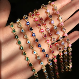 May include: A collection of gold-toned bracelets, each featuring a chain of heart-shaped charms. The charms are adorned with various colored gemstones and enamel, including green, blue, red, and pink. The bracelets are displayed against a dark background.