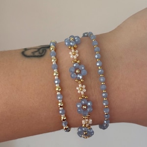 Può includere: Un set di tre braccialetti di perline con perline sfaccettate azzurre, piccole perline bianche a forma di fiore e perline distanziatrici color oro metallizzato. I braccialetti sono impilati, mostrando un mix di fili blu e un motivo floreale.