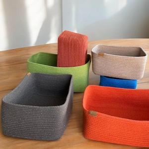 May include: A collection of woven storage baskets in various colors, including gray, green, orange, tan, and blue. The baskets are rectangular and have a textured appearance. Some baskets have a small leather tag attached.
