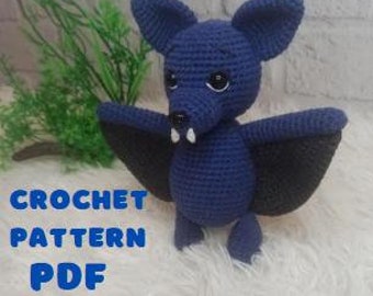 Pode incluir: Padrão de crochê PDF para um morcego amigurumi azul com asas pretas e dentes brancos. O morcego tem olhos pretos grandes e está em pé sobre uma superfície branca e felpuda.