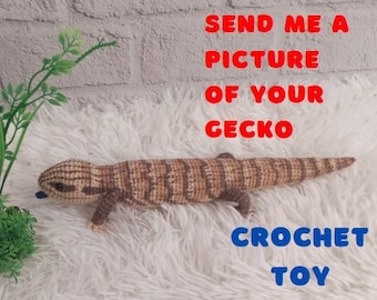 May include: Crocheted gecko toy with brown and white stripes. The toy is lying on a white fluffy surface. The text "SEND ME A PICTURE OF YOUR GECKO CROCHET TOY" is written in red and blue letters on a white background.