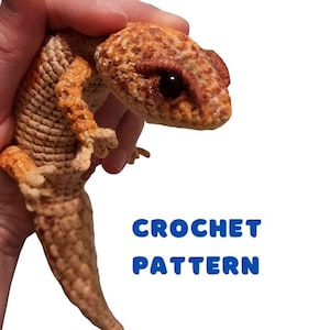 May include: Crocheted gecko toy in brown and orange colours. The toy is being held by a hand. The text "CROCHET PATTERN" is written in blue letters below the toy.