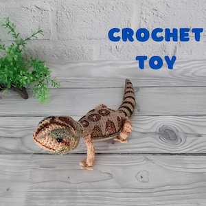Peut inclure: Lézard en crochet jouet avec des écailles brunes et beiges. Le lézard est posé sur une surface en bois clair. Le texte "CROCHET TOY" est en lettres bleues au-dessus du lézard.