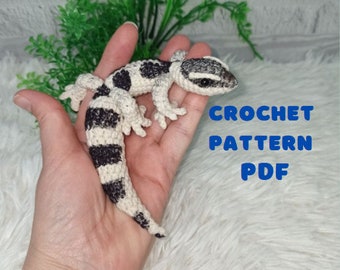 May include: Crocheted gecko amigurumi toy in white and black stripes. The toy is being held in a hand. The text "CROCHET PATTERN PDF" is visible in the image.