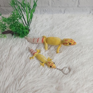 May include: Two handmade crochet geckos in yellow, orange, and white. One is a keychain, and the other is a larger decorative piece. The geckos have detailed patterns and are set against a fluffy white background with a green plant.