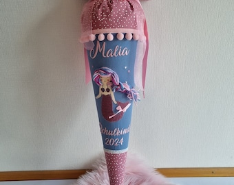 Puede incluir: Un cono de regalo de tela con cuerpo azul polvoriento, una aplicación de sirena, cabello de hilo rosa y el texto Malia Schulkind 2024. Las secciones superior e inferior son de tela rosa con puntos blancos, acentuadas con cinta y pompones.