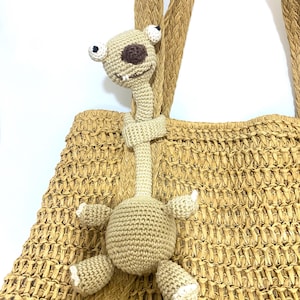 May include: A beige crocheted alien toy with large eyes, a long neck, and small arms and legs. The toy is attached to a woven straw tote bag with matching handles. The alien has a brown eye and white accents.