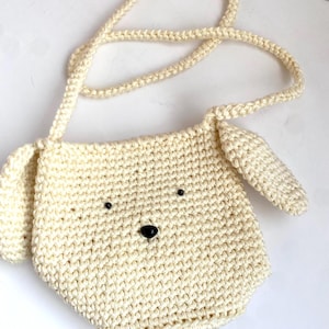 May include: Cream-colored crochet bag shaped like a dog's face, featuring black button eyes and a nose. The bag has long, floppy ears and a strap for carrying. The handmade bag is a unique accessory.