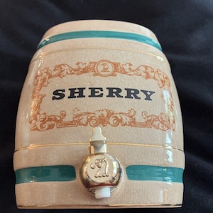 May include: A cream-colored ceramic barrel-shaped beverage dispenser with a crackle glaze finish. It features teal horizontal bands, gold trim, and an orange decorative frame surrounding the black text "SHERRY". A metallic spigot is attached.