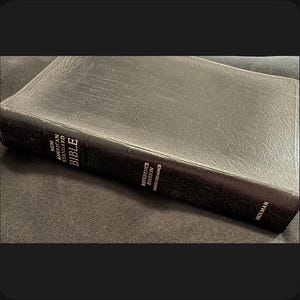 May include: A black leather-bound Bible with the text "New American Standard Bible" and "Reference Edition Concordance" visible on the spine. The book is closed and lies on a dark surface. The publisher's name, Holman, is also visible.
