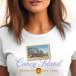 Può includere: Maglietta bianca con un'immagine di cartolina vintage del Cyclone, un ottovolante a Coney Island, Brooklyn, New York. Il testo "Coney Island" è stampato in un font stilizzato sotto l'immagine, con "Brooklyn" e "New York" stampati in un font più piccolo sotto il testo.