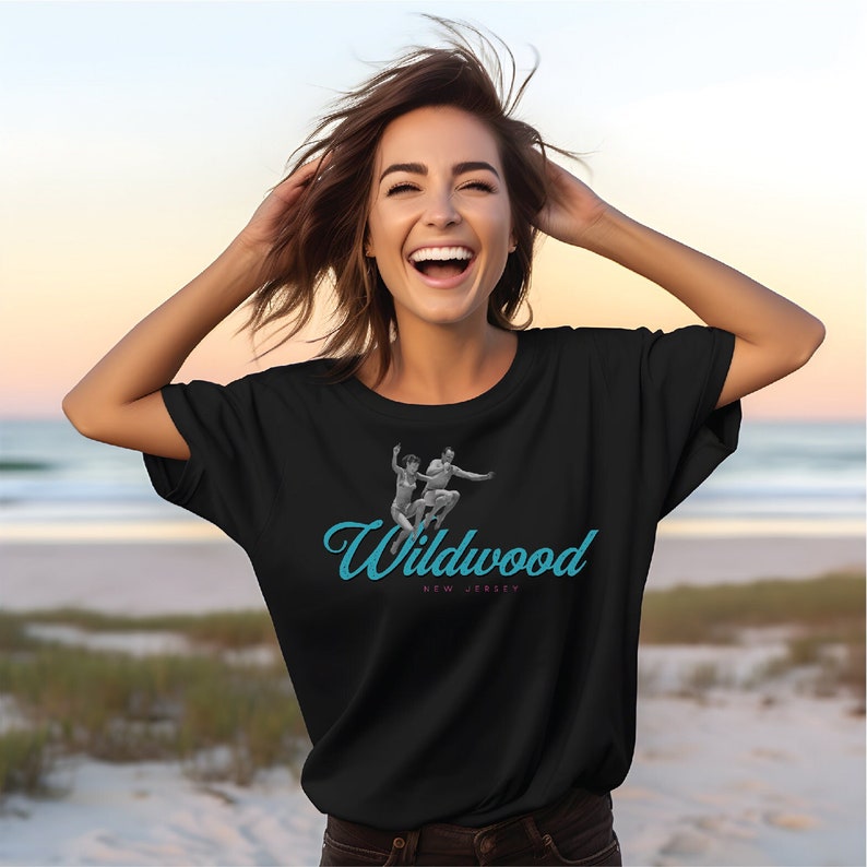 May include: A black t-shirt with a graphic of two people jumping in the air. The graphic is in black and white and the text "Wildwood NEW JERSEY" is in blue. The person wearing the shirt is smiling and has brown hair.