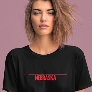 Puede incluir: Camiseta negra con la palabra "NEBRASKA" en rojo, centrada debajo de una línea horizontal roja. La camiseta la lleva una persona con el pelo corto y castaño claro y una expresión neutra. El fondo es de color morado apagado.