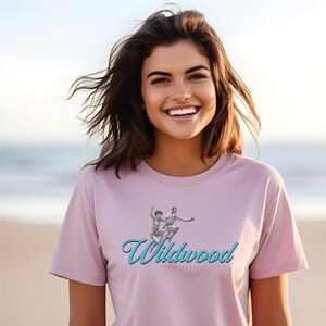May include: A pink t-shirt with a blue and white graphic of two people jumping in the air. The text "Wildwood New Jersey" is printed in blue below the graphic.