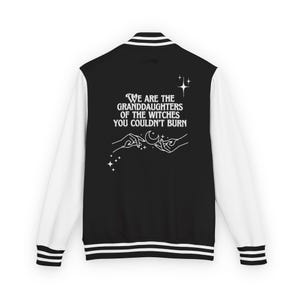 May include: Black and white letterman jacket with white sleeves. The back of the jacket features the text "WE ARE THE GRANDDAUGHTERS OF THE WITCHES YOU COULDN'T BURN" in white, along with a graphic of hands, a moon, and stars.
