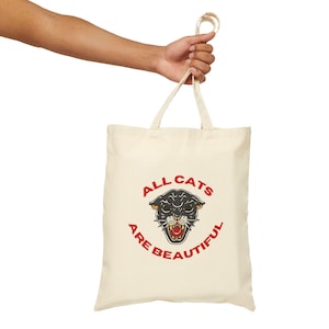 May include: A cream-coloured tote bag featuring the text "ALL CATS ARE BEAUTIFUL" in red and a black panther graphic. The bag is held by a person's hand. The tote bag has a simple design with long handles.