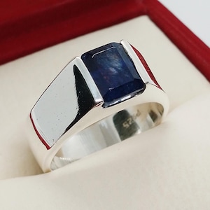 Op de afbeelding: Een zilveren ring met een donkerblauwe rechthoekige edelsteen. De ring ligt in een rode fluwelen doos.