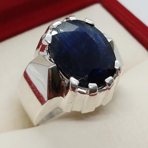 May include: A silver ring with a large, oval-shaped, dark blue gemstone set in a prong setting. The ring has a thick band with a textured design.