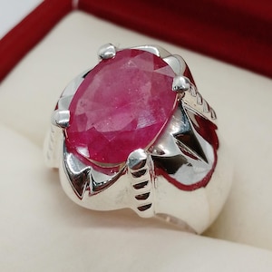 May include: A silver ring featuring a large, oval-shaped ruby gemstone. The ruby is a deep red colour and has a faceted cut. The ring has an elaborate design with intricate carvings.