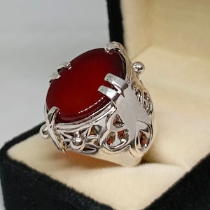 Könnte beinhalten: Ein silberner Ring mit einem großen, ovalen, roten Edelstein, der in einem filigranen Design gefasst ist.