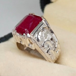 Peut inclure: Une bague en argent avec une grande pierre précieuse rouge carrée sertie au centre. La bande de la bague est finement gravée d'un motif floral.