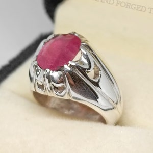 Peut inclure: Une bague en argent avec une grande pierre précieuse rouge de forme ovale sertie au centre. La bague présente un design détaillé et orné autour de la pierre précieuse.