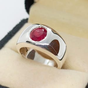 Peut inclure: Une bague en argent avec une grosse pierre précieuse rouge foncé de coupe ovale. La bague a un large anneau poli et un sertissage à lunette. La bague est présentée dans un écrin noir avec un intérieur beige.