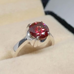 Peut inclure: Une bague en argent ornée d'une grosse pierre rouge ronde et facettée. La pierre est sertie dans un sertissage à quatre griffes, et le bracelet a un design simple et élégant. La bague est présentée sur une surface crème.