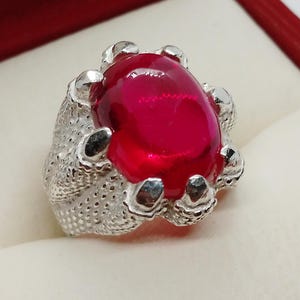 Pode incluir: Um anel de prata com uma grande gema vermelha ovalada engastada em uma garra. O anel tem um design detalhado com uma superfície texturizada.