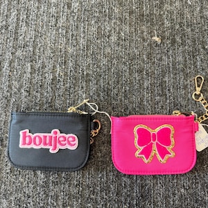 May include: Two small coin purses. One is black with the word "boujee" in pink. The other is hot pink with a gold glitter bow design. Both have gold-colored zippers and keychains.