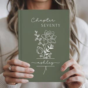 May include: A sage green journal with the words "Chapter Seventy" and "ashley" in white script. A floral line drawing is centered on the cover. The journal is held by a person.