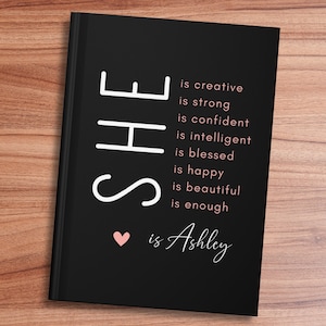 May include: Black journal with white text that reads "SHE is creative is strong is confident is intelligent is blessed is happy is beautiful is enough &hearts; is Ashley".