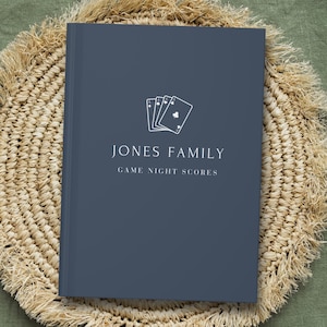 May include: A dark blue hardcover book with white text that reads JONES FAMILY GAME NIGHT SCORES and a graphic of four playing cards. The book is centered on a round, beige, woven straw placemat with a fringed edge.