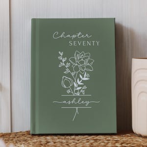 May include: A sage green hardcover journal with the words "Chapter Seventy" and the name "ashley" in white script. A floral line drawing is centered on the cover. The journal is displayed on a woven mat.