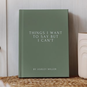 May include: A sage green hardcover book with the white text "THINGS I WANT TO SAY BUT I CAN'T" and "BY ASHLEY MILLER" printed on the front cover. The book is positioned upright, featuring a clean, minimalist design with centered typography.