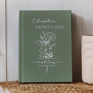 May include: A sage green hardcover journal with the text "Chapter Twenty-One" and the name "ashley" in white script. A floral line drawing is centered on the cover. The journal is displayed on a woven mat.