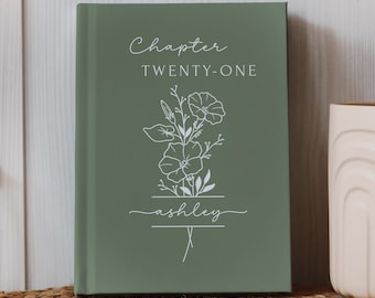 May include: A sage green hardcover journal with the text "Chapter Twenty-One" and the name "ashley" in white script. A floral line drawing is centered on the cover. The journal is displayed on a woven mat.