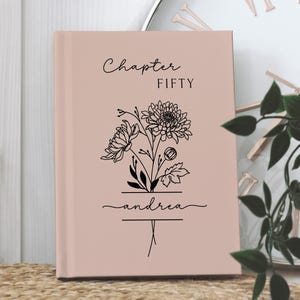 May include: A blush-colored journal with the words "Chapter FIFTY" and the name "andrea" in black script. A floral illustration is centered below the text. The journal is displayed on a woven surface.