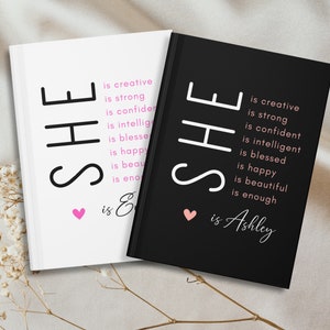 May include: Two black and white journals with the word "SHE" in large letters. The text "is creative, is strong, is confident, is intelligent, is blessed, is happy, is beautiful, is enough" is written in smaller letters below the word "SHE". The journal on the left has the text "is Enough" below the smaller text. The journal on the right has the text "is Ashley" below the smaller text. Both journals have a small pink heart above the text.