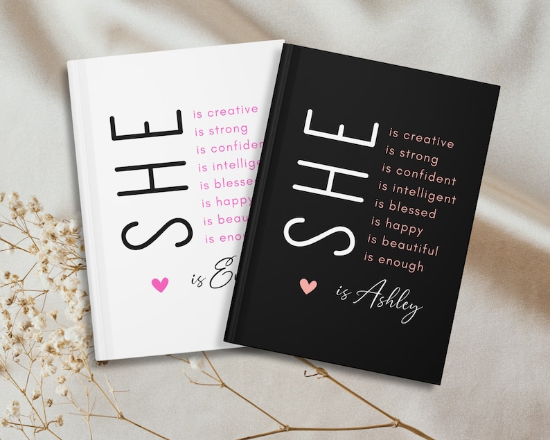 May include: Two black and white journals with the word "SHE" in large letters. The text "is creative, is strong, is confident, is intelligent, is blessed, is happy, is beautiful, is enough" is written in smaller letters below the word "SHE". The journal on the left has the text "is Enough" below the smaller text. The journal on the right has the text "is Ashley" below the smaller text. Both journals have a small pink heart above the text.