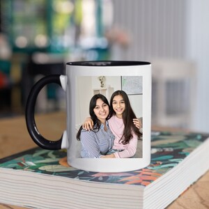 May include: White ceramic mug with a black handle and a black rim. The mug features a photo of two people smiling at the camera. The person on the left is wearing a striped shirt and the person on the right is wearing a pink shirt. The mug is sitting on a stack of books with a floral design.