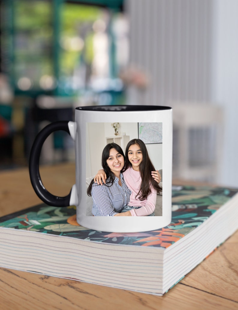 May include: White ceramic mug with a black handle and a black rim. The mug features a photo of two people smiling at the camera. The person on the left is wearing a striped shirt and the person on the right is wearing a pink shirt. The mug is sitting on a stack of books with a floral design.